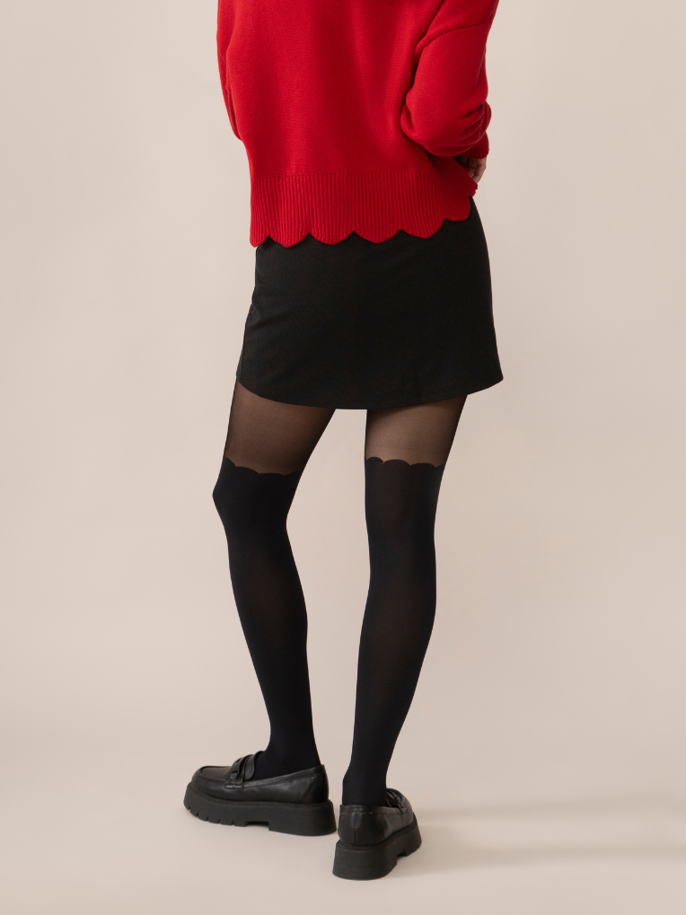 A person wearing over-the-knee black tights with a scalloped finish, paired with a red skirt and black shoes.