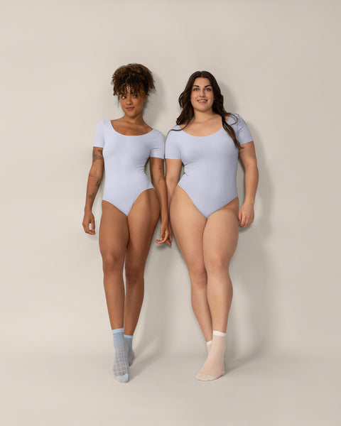 Two women wearing light blue bodysuits against a beige background