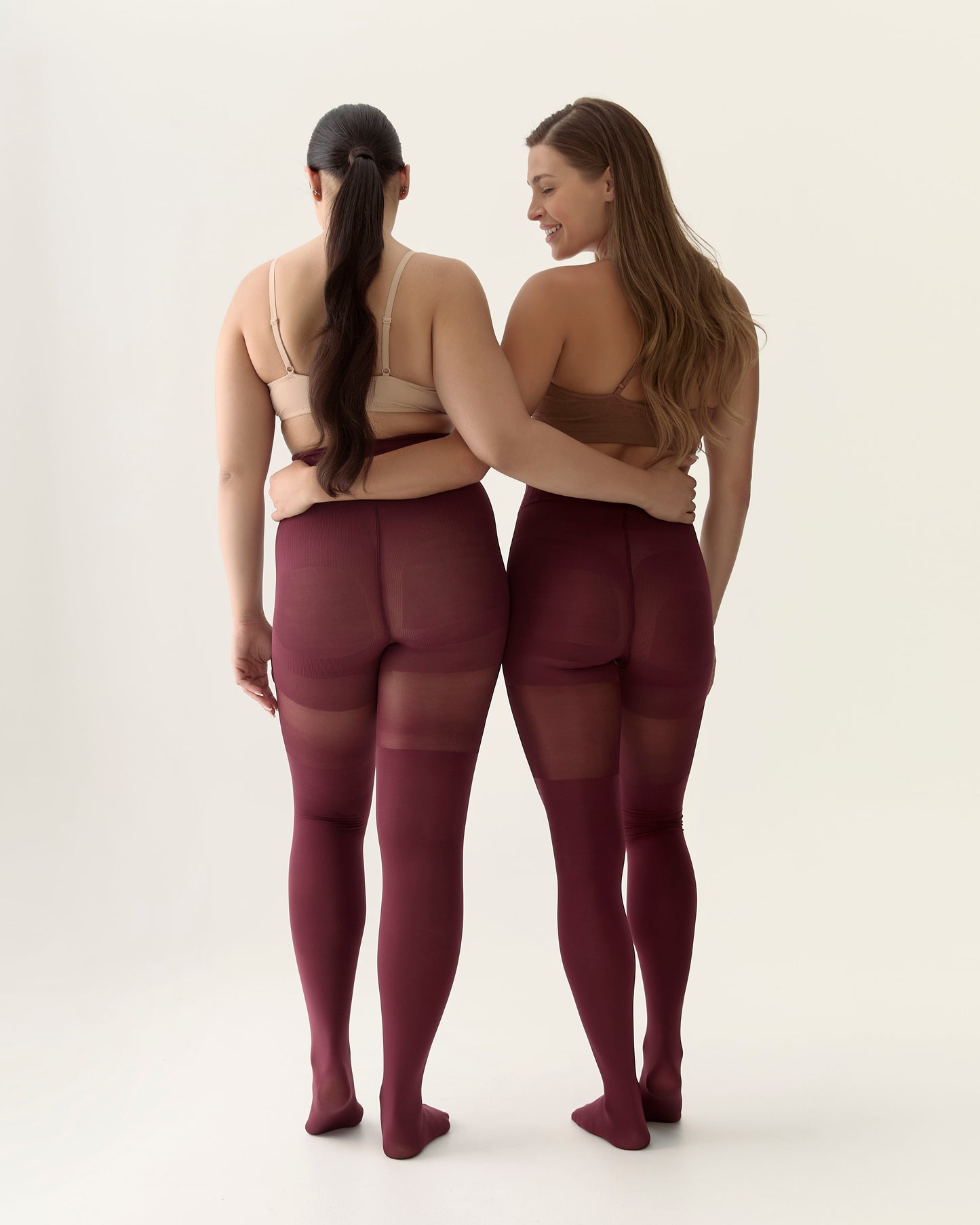 Two women wearing burgundy shaping tights and beige bras against a white background