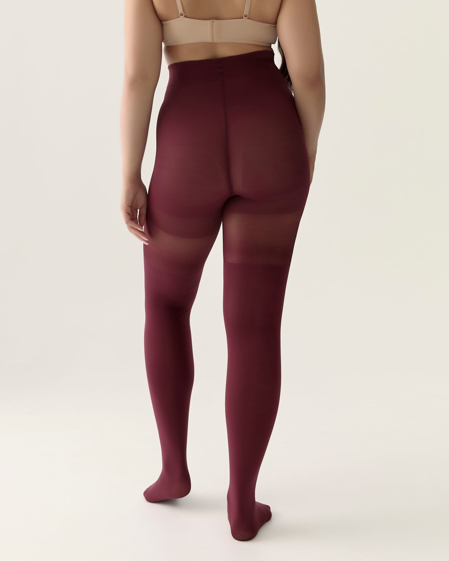 Person wearing a beige bra and burgundy shapewear tights on a plain background