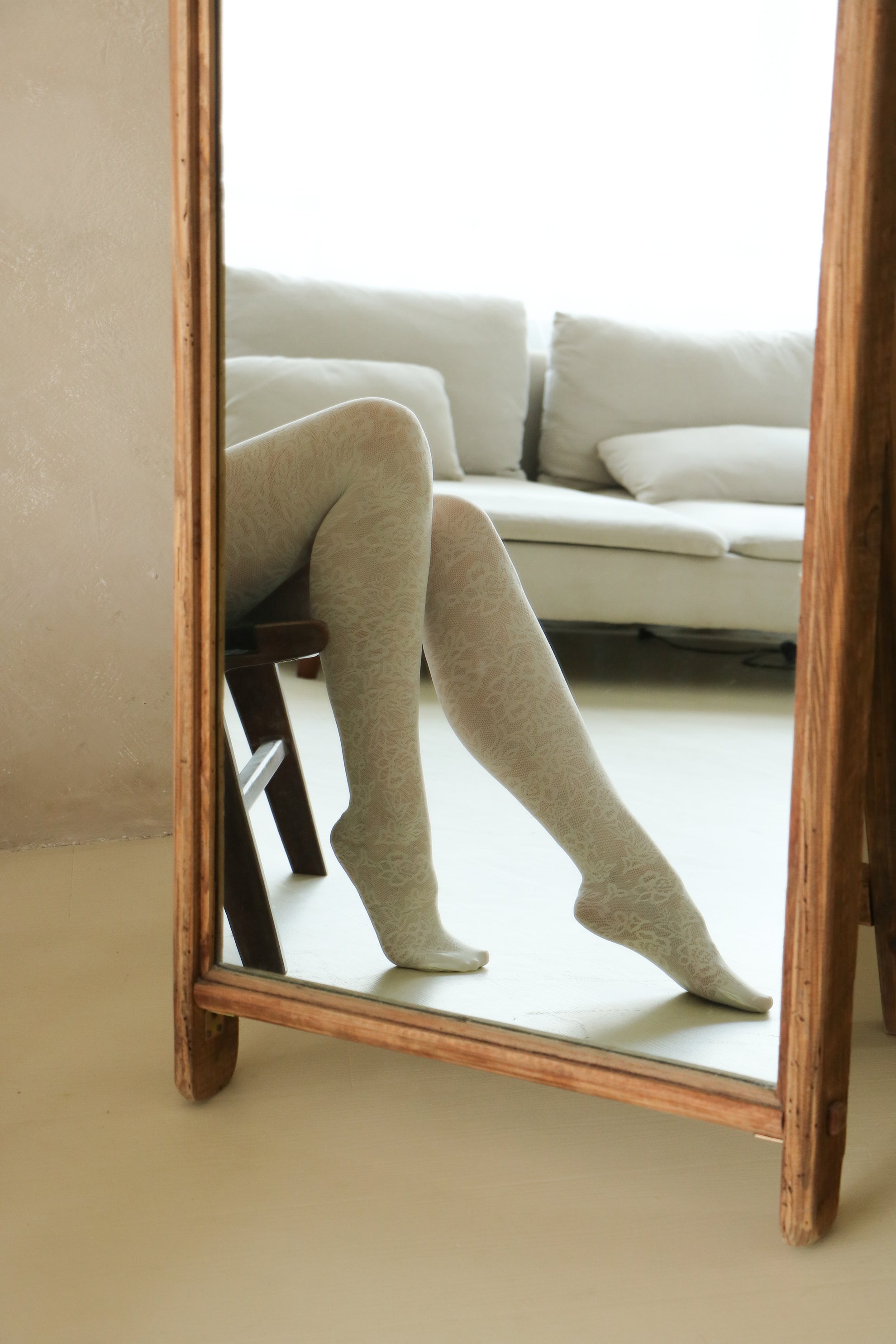 Person wearing beige tights and shoes reflected in a wooden-framed mirror.