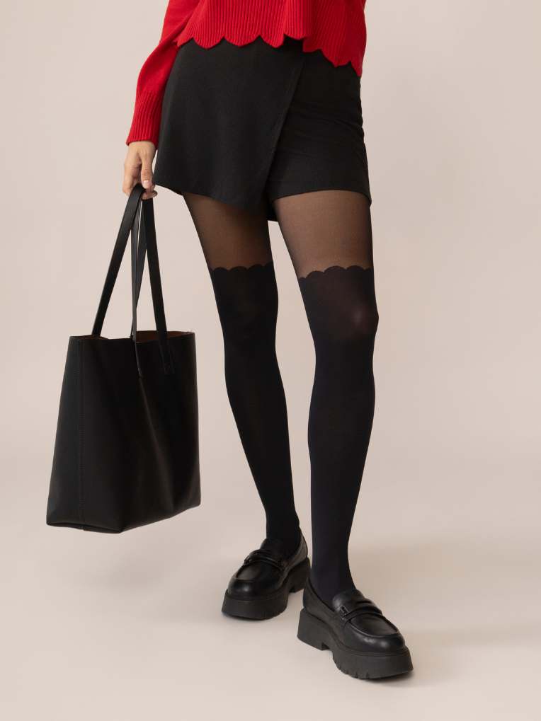 Person wearing black tights with scallop details, holding a black tote bag on a beige background