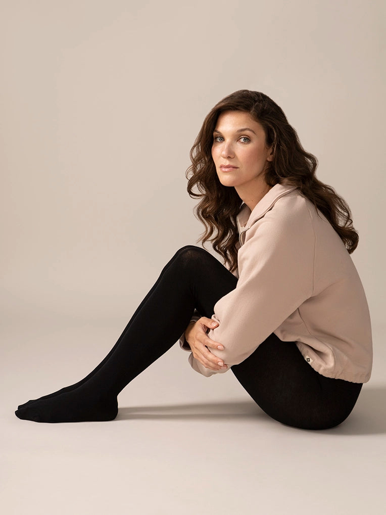 Woman sitting on the floor wearing black tights and a beige jacket against a plain background